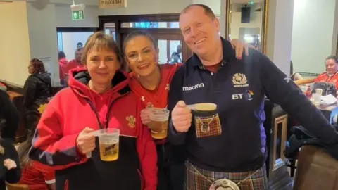 Francesca Murphy Francesca stands in the pub between her mother and father. They are all smiling and looking happy and holding pints of beer.