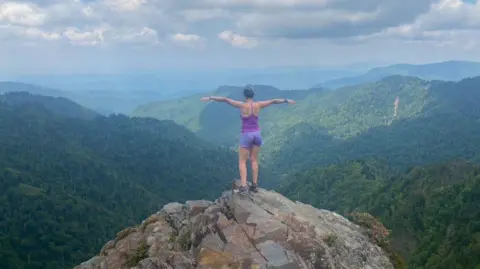 Francesca Murphy Francesca Murphy stands on top of a hill with a beautiful view of the green, lush mountains. Her back is to the camera and she stretches out her arms and looks at the view. 