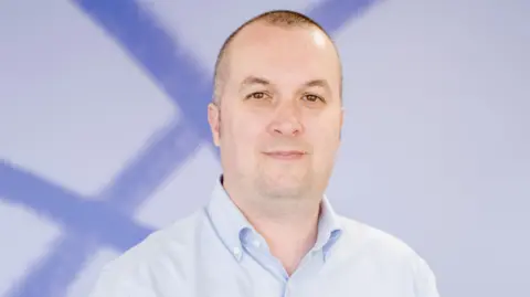 Mind Cymru Simon Jones, a white man with shaved brown/grey hair looks into the camera with a slight smile. He wears a light blue shirt and stands in front of a white and blue background.